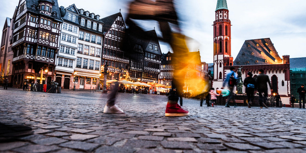 [Translate to English:] Blick auf einen Marktplatz einer Stadt mit vielen alten Gebäuden. Es sind verschwommene Umrisse im Bild zu sehen von einer Person die gerade vorbeigeht.