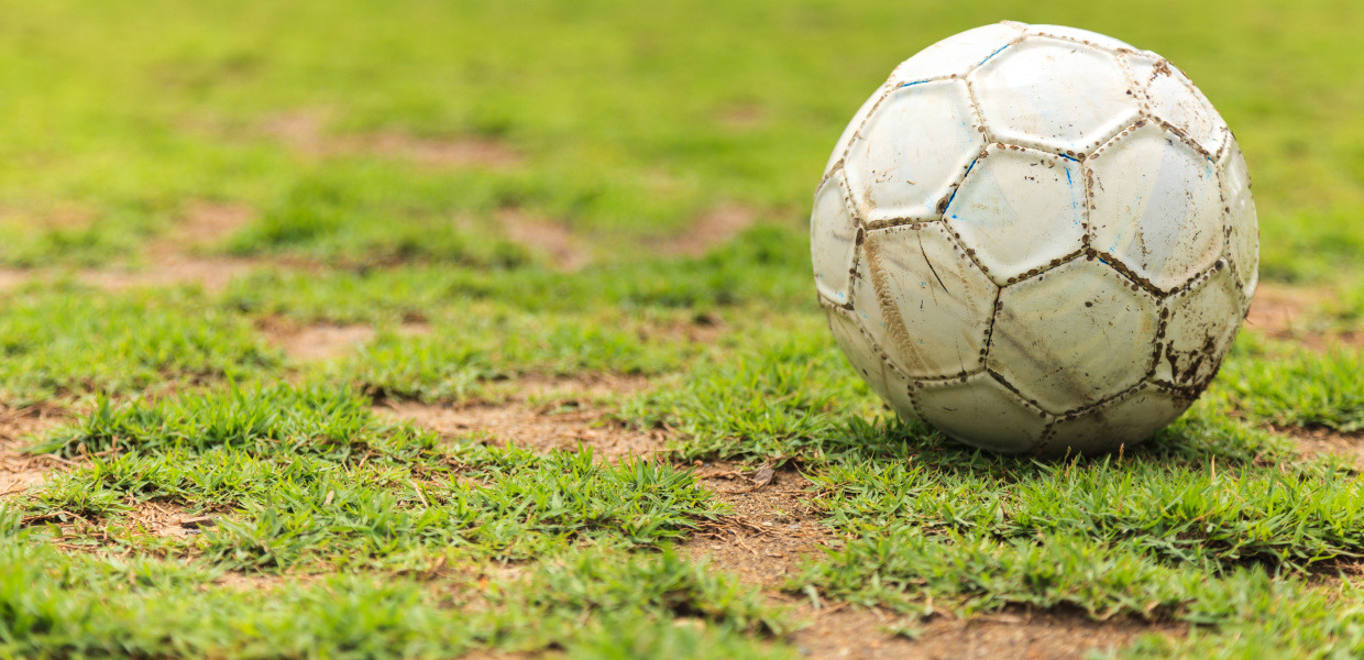 (© naypong - stock.adobe.com) Ein alter, abgenutzter Fußball mit Erd- und Farbspuren liegt auf einem löchrigen Fußballrasen.