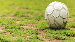 Ein alter, abgenutzter Fußball mit Erd- und Farbspuren liegt auf einem löchrigen Fußballrasen.