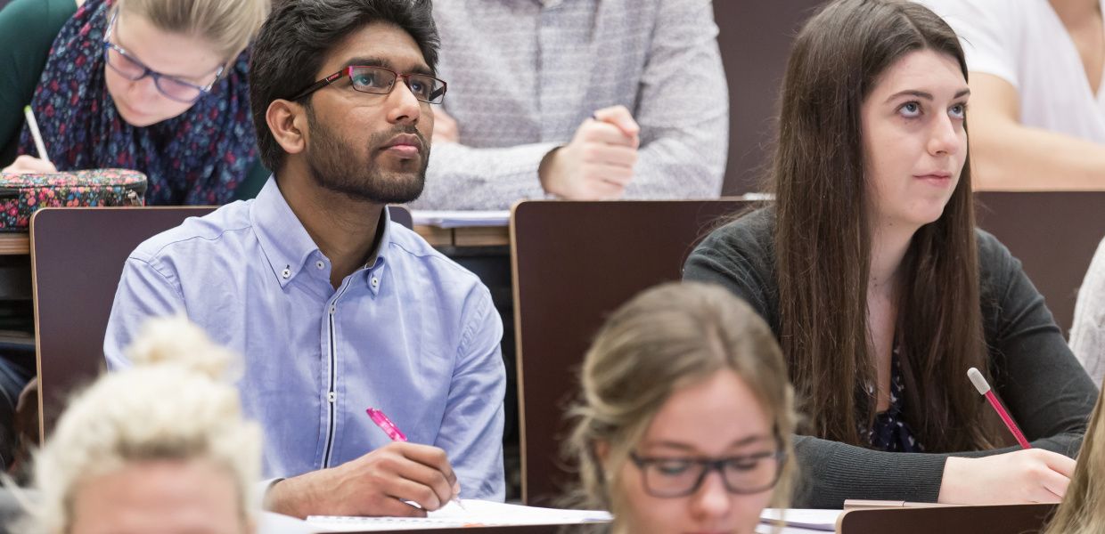 (© Universität Wien / Flickr - CC BY-NC 2.0 https://creativecommons.org/licenses/by-nc/2.0/) Junge Menschen sitzen in einem Hörsaal. Einige lauschen einem Vortrag und blicken auf, andere schreiben.
