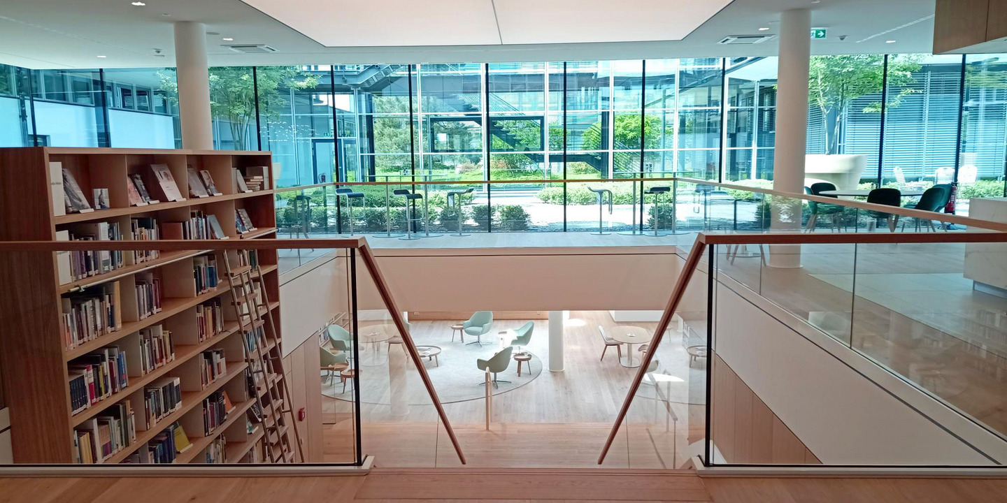 Blick vom oberen Bereich des Workcafés im Gütersloher Gebäude der Bertelsmann Stiftung in den unteren Bereich mit der Stiftungsbibliothek. Vom oberen Bereich führt eine Treppe in den unteren Bereich, daneben steht ein Hochregal mit Büchern. Im Hintergrund ist ein begrünter Innenhof zu sehen.