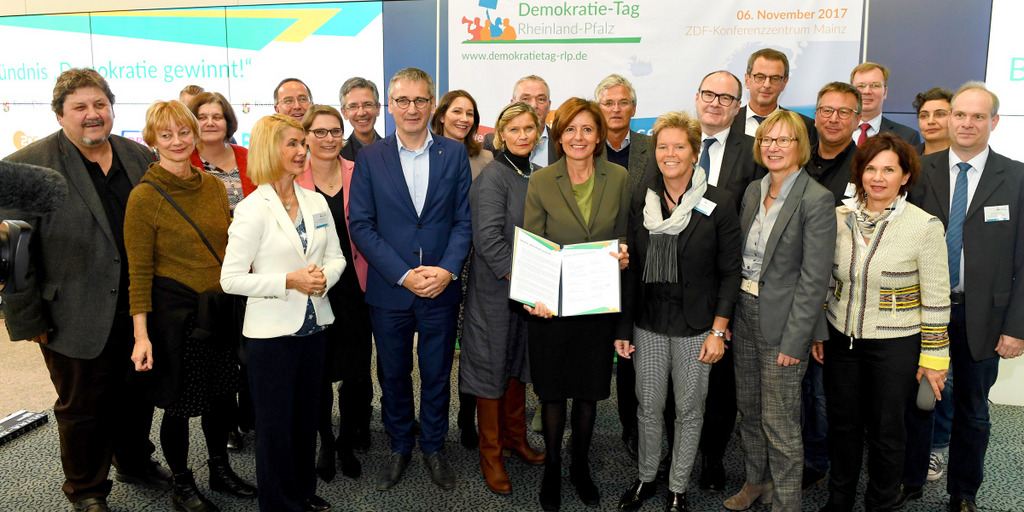 Die Ministerpräsidentin von Rheinland-Pfalz, Malu Dreyer, hält das Gründungsdokument für das Bündnis "Demokratie gewinnt!" in die Kamera und lächelt. Dabei steht sie umgeben von Vertretern der Bündnispartner, darunter unser Vorstandsmitglied Brigitte Mohn.