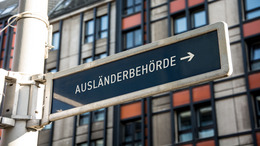 Im Hintergrund ist ein Gebäude zu sehen, im Vordergrund ein Schild mit der Aufschrift "Ausländerbehörde"