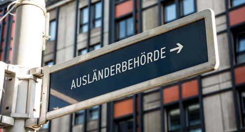 Im Hintergrund ist ein Gebäude zu sehen, im Vordergrund ein Schild mit der Aufschrift "Ausländerbehörde"