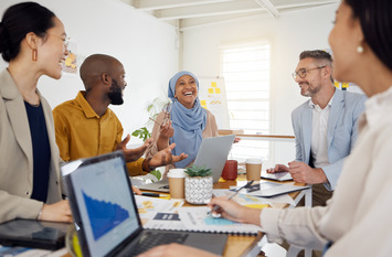 Ein Team aus verschiedenen Mitarbeitenden, darunter eine Frau mit Kopftuch und ein Mann mit dunkler Hautfarbe, sitzen zusammen an einem Besprechungstisch und lachen.