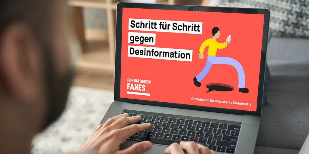Ein Mann tippt auf der Tastatur seines Laptops, auf dem ein Bild mit dem Logo des "Forum gegen Fakes" sowie dem Text "Schritt für Schritt gegen Desinformation" zu sehen ist.