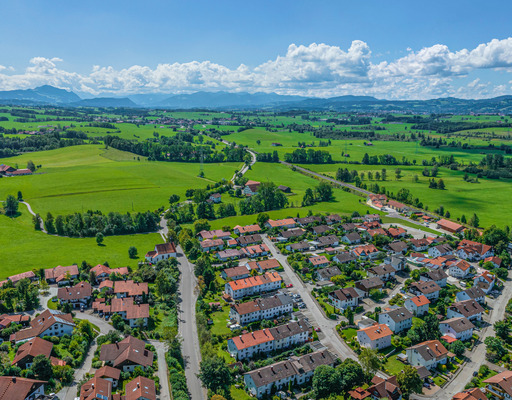 Das Bild zeigt eine Panoramaaufnahme des bayerischen Energiedorfs Wildpoldsried. Eingebettet in eine grüne Hügellandschaft mit Blick auf die Alpen im Hintergrund, präsentiert sich der Ort mit zahlreichen Einfamilienhäusern, vielen Photovoltaikanlagen auf den Dächern und einer klaren, strukturierten Bebauung.