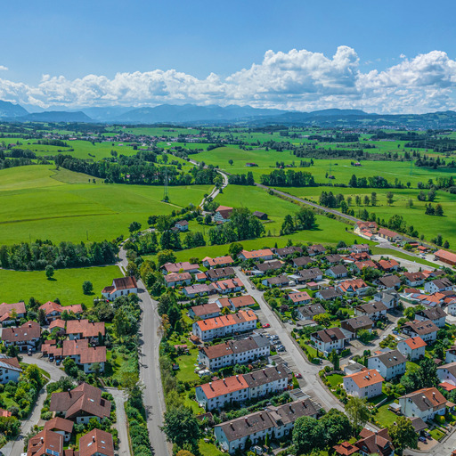 Das Bild zeigt eine Panoramaaufnahme des bayerischen Energiedorfs Wildpoldsried. Eingebettet in eine grüne Hügellandschaft mit Blick auf die Alpen im Hintergrund, präsentiert sich der Ort mit zahlreichen Einfamilienhäusern, vielen Photovoltaikanlagen auf den Dächern und einer klaren, strukturierten Bebauung.
