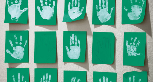 An der Wand einer Kita hängen grüne Blätter Papier, auf denen Kinder in weißer Farbe ihre Handabdrücke hinterlassen haben.