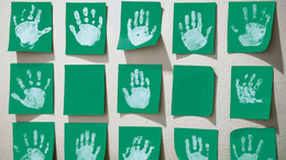 An der Wand einer Kita hängen grüne Blätter Papier, auf denen Kinder in weißer Farbe ihre Handabdrücke hinterlassen haben.