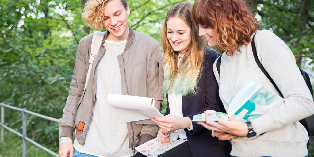 Drei junge Menschen schauen gemeinsam auf einen Papierblock. Dabei befinden sie sich draußen vor einer mit Bäumen bewachsenen Fläche.