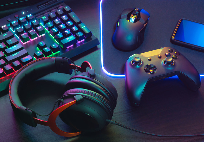 Das Bild zeigt ein Set aus Gaming-Zubeh&ouml;r auf einem Schreibtisch: eine beleuchtete mechanische Tastatur, eine Gaming-Maus auf einem leuchtenden Mauspad, ein schwarzer Gamecontroller, ein Smartphone und ein Paar Over-Ear-Kopfh&ouml;rer. Alle Ger&auml;te sind in farbigem RGB-Licht inszeniert.