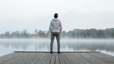 Young man standing alone on wooden footbridge and staring at lake. Thinking about life. Mist over water. Foggy air. Early chilly morning. Peaceful atmosphere in nature. Enjoying fresh air. Back view.(© fotoduets - stock.adobe.com)