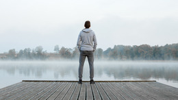 Ein Mann steht auf einem Steg an einem See, dessen Wasseroberfläche leicht im Nebel liegt. Der Mann ist von hinten zu sehen, wie er über den See hinweg in die Ferne blickt.