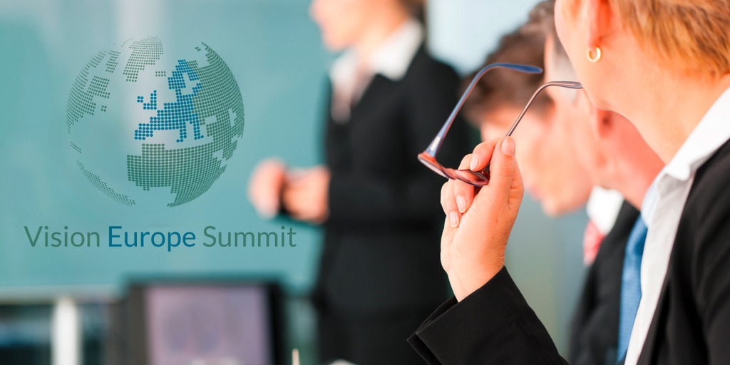 Four people in a room: A woman is standing beside a wall, where the words "Vision Europe Summit" and a globe can be seen. Another woman and two men are sitting in on a table looking at her.