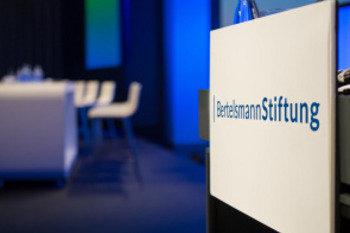 Zu sehen ist ein Stehpult mit dem Schriftzug Bertelsmann Stiftung. Im Hintergrund steht ein Tisch mit Stühlen.
