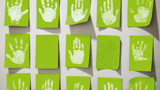 Hellgrüne Post-It-Zettel kleben an einer Wand. Auf einigen von ihnen sind Abdrücke von Kinderhänden zu sehen.