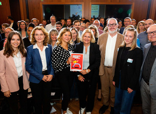 Gruppenfoto von Bundesinnenministerin Nancy Faeser, Daniela Schwarzer, Vorständin der Bertelsmann Stiftung, und Teilnehmenden des Bürgerrates bei der Übergabe des Bürgergutachtens zum "Forum gegen Fakes". Nancy Faeser und eine Teilnehmende des Bürgerrates werden von den anderen Teilnehmenden umgeben und halten lächelnd das Bürgergutachten in die Kamera.