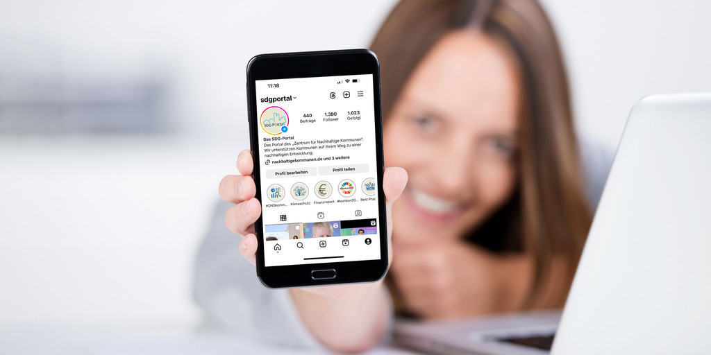 Eine Frau hält ein Handy weit nach vorn. Auf dem Handydisplay ist die Profilseite des Instagram-Kanals sdgportal zu sehen. Die Frau im Hintergrund ist unscharf dargestellt.