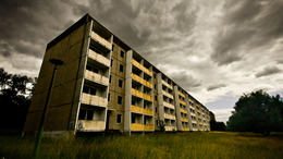 Ein verfallenes Hochhaus in dunklen und dreckigen Farben. Das Foto ist sehr düster und dunkel. Am Himmel ziehen graue Wolken.