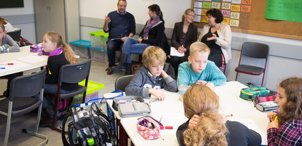 Kinder und Lehrer sitzen locker verstreut im Klassenraum und sprechen miteinander