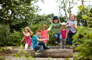 Eine Gruppe fröhlicher Schulkinder rennt durch einen Garten oder Park und springt über einen Holzbalken.