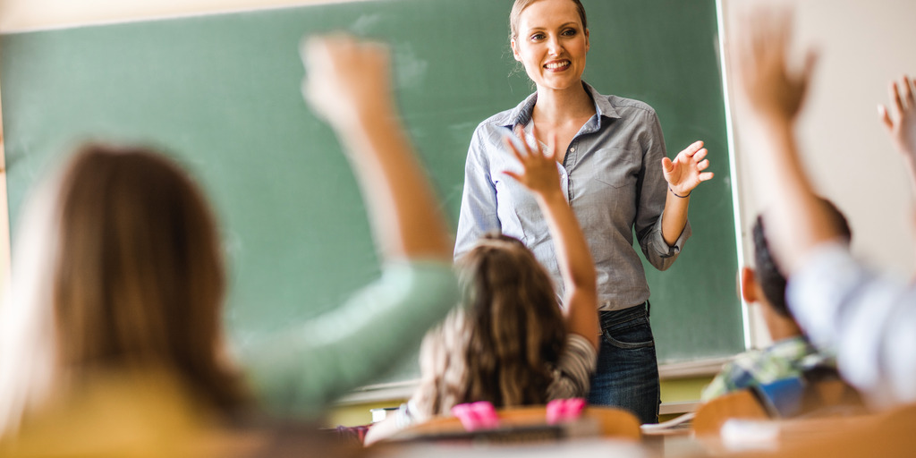 Eine Lehrerin steht an der Tafel und stellt den Schüler:innen eine Frage