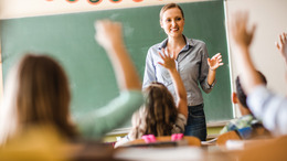 Eine Lehrerin steht an der Tafel und stellt den Schüler:innen eine Frage