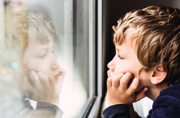 Ein Junge steht vor einem Fenster. Er stützt die Ellenbogen auf und hält mit den Händen seinen Kopf.