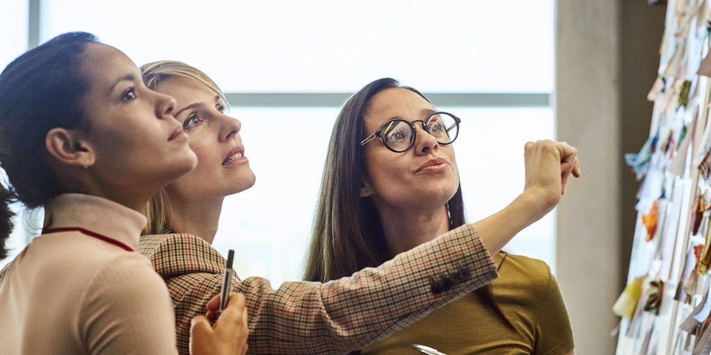 Drei Frauen stehen an einer Metaplanwand. Eine dunkelhaarige ist von der Seite zu sehen, neben ihr eine  blonde zeigt auf etwas an der Wand und die dritte daneben ist von vorn zu sehen. Alle drei schauen auf die Wand und die mittlere Frau zeigt auf etwas an der Wand.