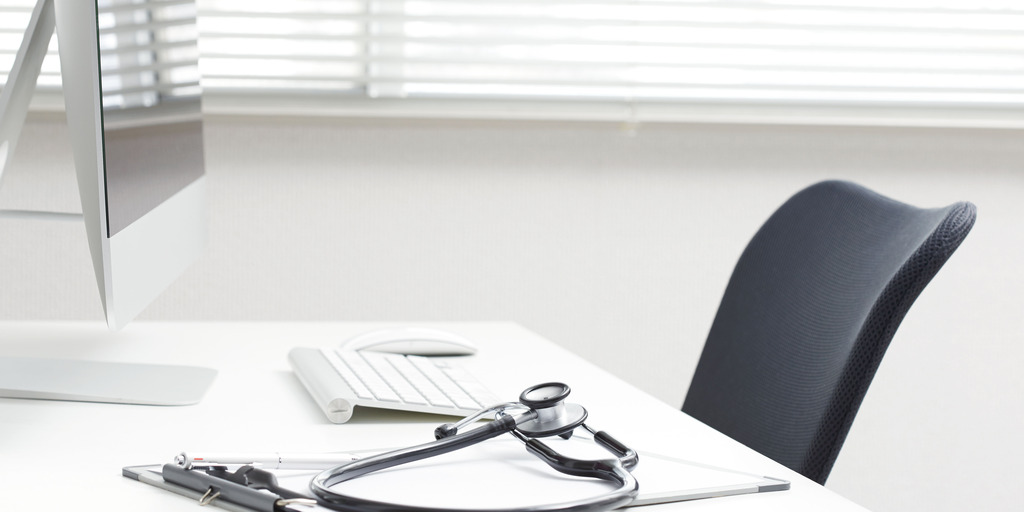 Das Bild zeigt einen hellen, modernen Schreibtisch in einer Arztpraxis oder einem medizinischen Büro. Auf dem Tisch liegen ein Stethoskop, ein Klemmbrett und ein Stift. Im Hintergrund sind ein Computer mit Tastatur und Maus sowie ein ergonomischer Stuhl zu sehen.