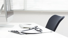 Das Bild zeigt einen hellen, modernen Schreibtisch in einer Arztpraxis oder einem medizinischen Büro. Auf dem Tisch liegen ein Stethoskop, ein Klemmbrett und ein Stift. Im Hintergrund sind ein Computer mit Tastatur und Maus sowie ein ergonomischer Stuhl zu sehen.