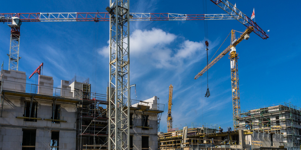 Der Blick zeigt eine Baustelle. Vor und hinter den neuen mehrstöckigen Häusern stehen Kräne. Der Himmel ist blau.