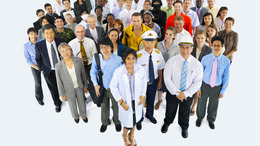 A group of people in working dress, including a doctor, a pilot, a firefighter, business people and construction engineers. They are standing closely together in a large group and smile into the camera.