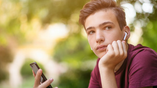 A teenage boy rests his head on his left hand and looks into the distance, in a pensive mood. In his right hand, he holds his smartphone.