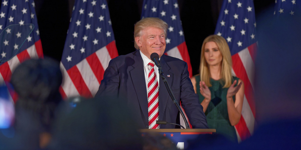 Donald Trump gives a campaign speech in September 2016 in Aston, Pennsylvania. He stands behind the lectern, smiling. His daughter Ivanka applauds in the background, standing before a row of American flags.