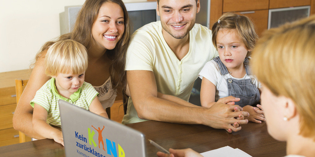 Eine Familie sitzt an einem Tisch und wird beraten. Auf dem Tisch steht ein Laptop mit dem Logo des Projekts Kein Kind zurücklassen.