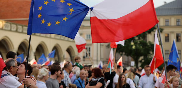 A large group of people with flags in their hands demonstrate in front of a historic building. In the foreground, one can see a European and a Polish flag tied together.