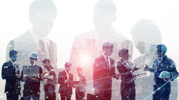 Business people holding laptops and tablets in their hands and engineers wearing construction helmets are shown standing in a row and talking to each other in small groups.