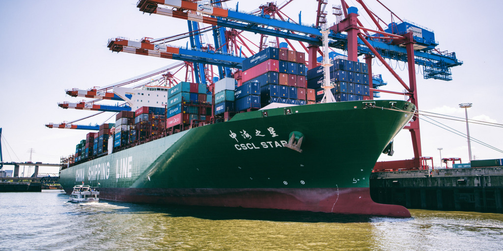 A Chinese container vessel anchors in Hamburg harbor. From the right side cranes are overlapping the ship.