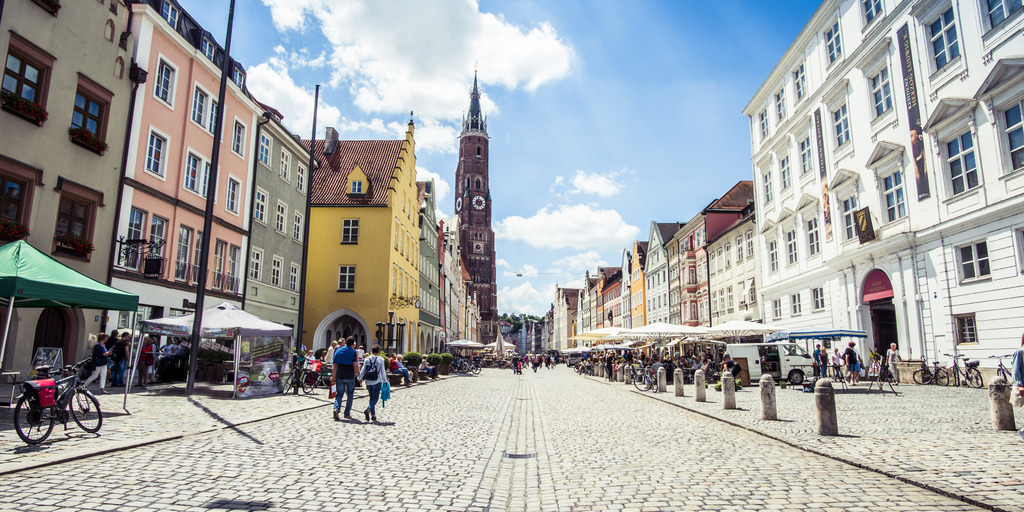 Auf dem Foto ist eine Fußgängerzone zu sehen. Links und rechts sind Geschäfte und Lokalitäten. Vor den Lokalitäten stehen Sonnenschirme. Der Himmel ist blau mit kleinen Wolken.