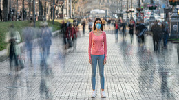 Eine junge Frau, die einen Mund-Nasen-Schutz trägt, steht in einer Großstadt auf einem belebten Bürgersteig und blickt in die Kamera, während um sie herum die anderen Passanten nur als flüchtige Schatten zu erkennen sind.