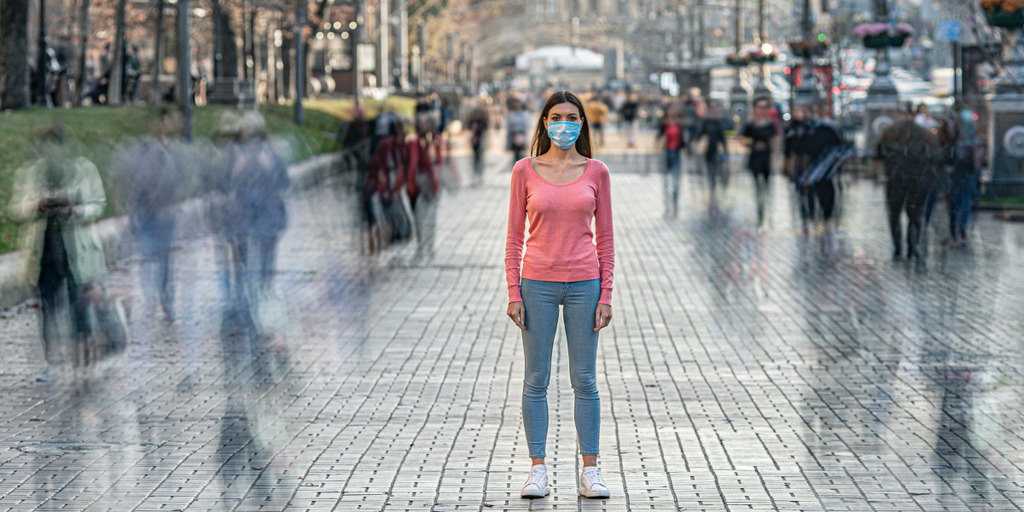 Eine junge Frau, die einen Mund-Nasen-Schutz trägt, steht in einer Großstadt auf einem belebten Bürgersteig und blickt in die Kamera, während um sie herum die anderen Passanten nur als flüchtige Schatten zu erkennen sind.
