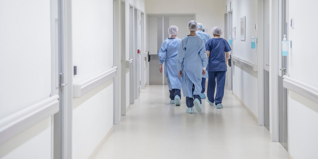 Vier Ärzte im Kittel und mit Haarschutz laufen einen Gang in einem Krankenhaus entlang. Man sieht sie nur von hinten.