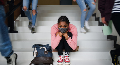Eine junge Studentin mit Gesichtsmaske zum Schutz vor dem Corona-Virus sitzt auf Treppen eines Campus . Sie wirkt nachdenklich, fast depressiv.