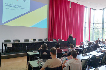 In einem Konferenzraum sitzen viele Personen. Sie sitzen vor ihren Laptops. Im Hintergrund ist eine Leinwand zu sehen.