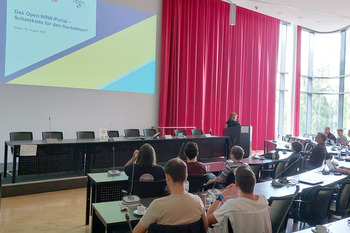 In einem Konferenzraum sitzen viele Personen. Sie sitzen vor ihren Laptops. Im Hintergrund ist eine Leinwand zu sehen.