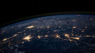 The Earth viewed from space at night. One can see clouds, some brightly lit cities and the curvature of the globe.