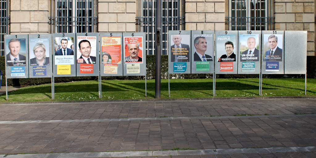 Plakatständer aus Metall, beklebt mit 11 Plakaten, stecken im Rasen vor einem Gebäude. Auf den Plakaten sind die Kandidaten für die französische Präsidentschaftswahl 2017 zu sehen.