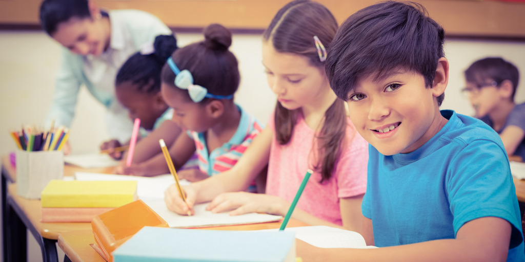 Grundschulkinder sitzen an ihren Tischen und lernen. Die Lehrerin steht am Ende der Reihe und beugt sich zur Hilfe über eine Schülerin.